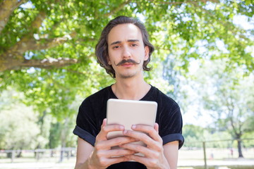 Front view of serious man with tablet looking at camera. Thoughtful young brunet with tablet posing in park. Technology concept