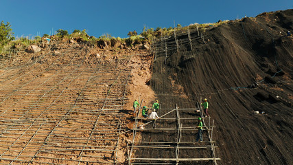 Protection of road from mountain slough, rockfall with metal accumulative restraining net fences. Workers constructing anti-landslide concrete wall prevent protect against rock slides. Rockfall