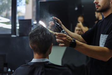 Attractive male is getting a modern haircut in barber shop. Barber wets hair by spray and combs them.