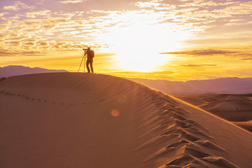 Photographer behind tripod standing with camera on sand dune silhouetted by sunrise in desert, Death Valley California