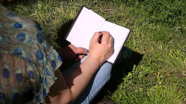 Woman and student sitting on grass, takes notes in notebook, learning and writes thoughts, writes book, she is preparing his final exams doing homework and thinking, writing something in his notebook.
