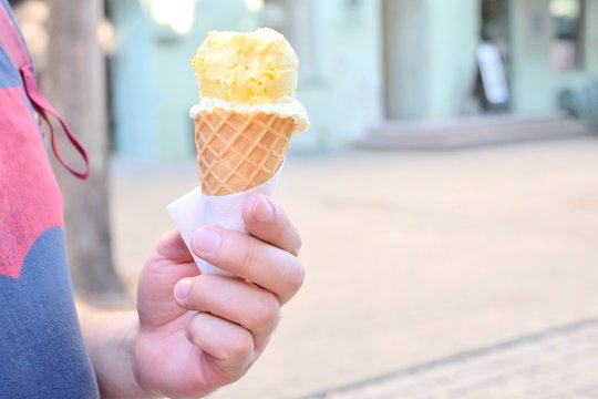 Man Holding Delicious Mango Ice Cream In Waffle Cone With Selective Focus On Blurred City Background. Frozen Summer Snack. Refreshing Lemon Icecream In The Man Hand. Cold Tasty Vanilla Ice Creams 