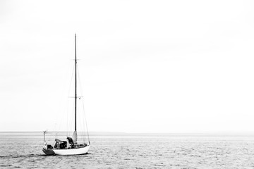 Lonely small yacht goes to the open sea. The man on the yacht is doing something. Grey sky. The sea is calm. There is a place for text. Black and white photo.
