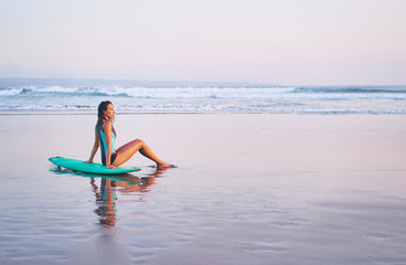 Surfing and vacation. Holiday on the beach. Relaxed young woman siiting on the sand with surf board enjoying sea view.