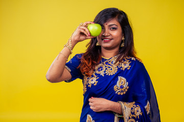 Close up portrait of a young Indian brunette woman with traditional blue sari in studio in yellow copy space background biting an apple in her hand