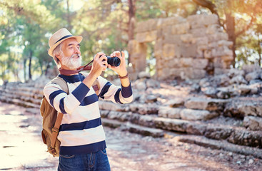 Enjoying travel. Senior man with backpack taking photo on camera on ancient sightseeing background.