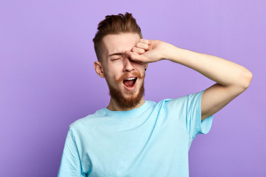 Handsome Bored Sleepy Bearded Man Touching His Eyes After Long Overwork On New Project, Wants To Sleep In The Morning, Close Up Portrait. Isolated Blue Background, Studio Shot