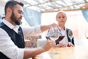 Young bearded male sommelier or bartender pouring red cabernet into wineglass