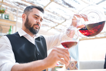 Young bearded barman or sommelier pouring red wine into wineglass