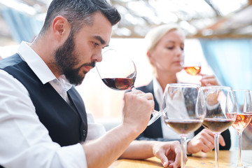 Young sommelier feeling smell of red wine while holding wineglass by his nose - obrazy, fototapety, plakaty