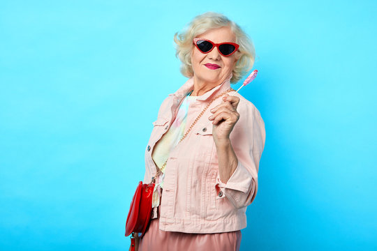 Old cheerful happy lady with sunglasses holding lollipop and looking at the camera on a blue background. sweets, love, valentine's day. fashion concept