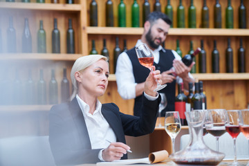 Blonde female sommelier examining sample of wine or brandy in bokal