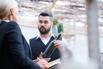 Young confident cavist with bottle of wine talking to his colleague
