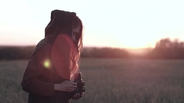 Photographer girl stands in field with vintage camera, on background sunset with sun glare.