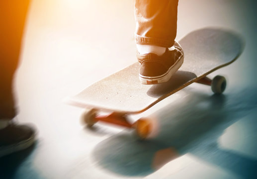 The guy in jeans and sneakers quickly pushes his foot off the asphalt and rides a skateboard, illuminated by bright sunlight. The picture shows the speed and dynamics of what is happening.