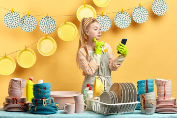 charming beautiful girl in gloves holding smart phone while doing dishes in modern light yellow...