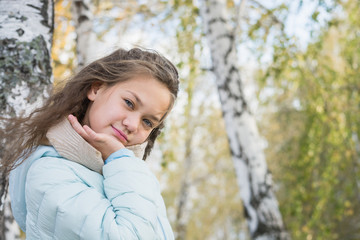 Beautiful portrait of young girl with long hair in blue down jacket at birch on an autumn day.