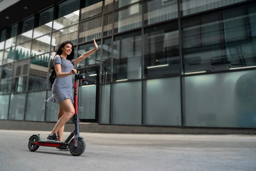 Cute young woman waving while standing on electric scooter 