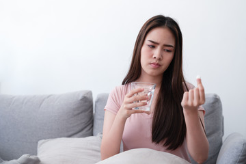 Unhappy sick asian young woman holding drug and glass of water painkiller to relieve abdominal pain while sitting on sofa couch in living room at home.