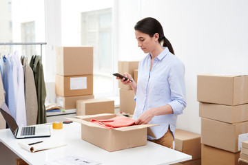 Young mobile female with smartphone taking photo of folded pullover in open box