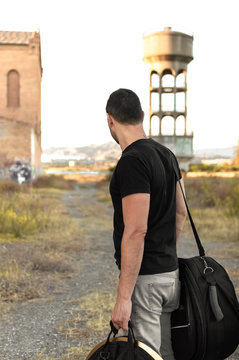 Man with casual clothes and luggage lost in the outskirts of the city at sunset. Exploring countryside and abandoned buildings in the urban suburbs.