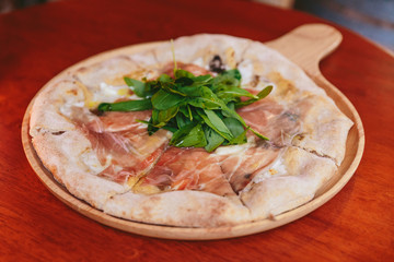 Parma Ham Pizza topping with rocket on rounded wooden plate with sliced bread in wooden box in the background.