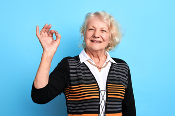 Smiling cheerful senior lady gesturing, showing ok sign. isolated blue background. studio shot....