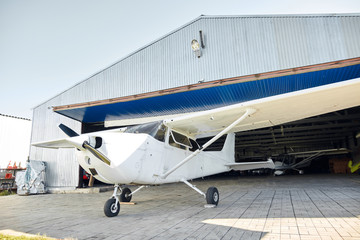 white single-engine airplane on three wheels stands before small aircraft hangar building, prepares to the flight, parked and ready to take off