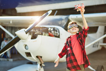 happy little boy playing with small toy airplane near hangar and white propeller air jet, wears...