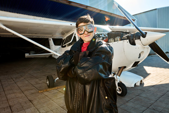 portrait of sweet little boy stands proudly in his father's black leather pilot jacket, wears aviator glasses, looking with smile in camera, white propeller aircraft stands on the background.