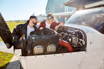 dad in pilot jacket and his kid aviator glasses inspecting small propeller airplane that is parked outside aircraft hangar building, near the airfield.
