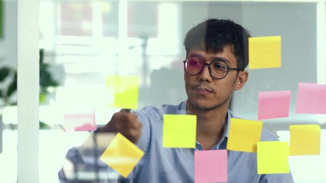 Asian creative man stick a sticky note on glass board. Young professional business male write information, strategy, reminder on paper, business situation, startup in Loft office concept.