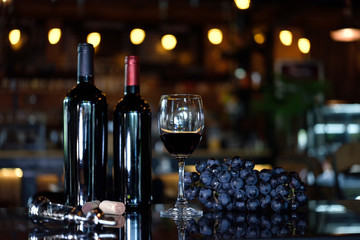 A glass of wine placed on the floor with 2 bottles of red wine and red grapes placed next to it