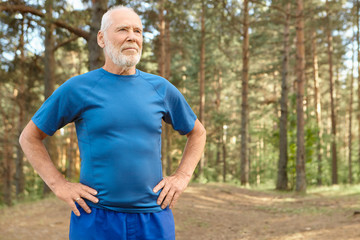 Naklejka na ściany i meble Self determined elderly man on retirement training outdoors in pine wood, holding hands on his waist, doing exercises to warm up body before run. Bearded retired male catching breath after workout