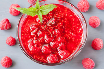 Glass small saucer with raspberry jam with fresh raspberries and mint close-up on gray background.