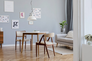 Design scandinavian home interior of open space with stylish chairs, family table wooden commode, gray sofa, accessories and mock up posters gallery wall. Gray background walls. Retro cozy home decor.