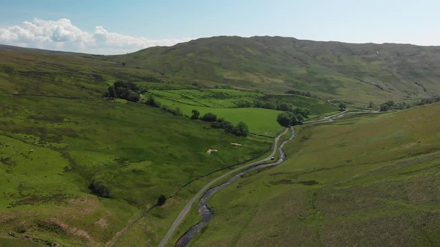 A high-altitude aerial shot of Barbon Beck, Carnforth, Lancashire, England. The camera moves slowly into the valley. This countryside footage features a stream running alongside a small country lane.