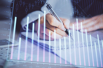A woman hands writing information about stock market in notepad. Forex chart holograms in front....