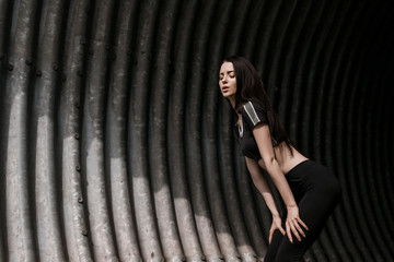Sports woman rest after run in fashion sportswear on tunnel urban gray background. Fitness model working out outdoor. Young beautiful slim brunette girl in trendy black leggings and top.