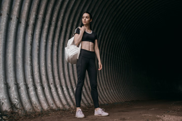 Sports woman posing in fashion sportswear on tunnel urban gray background. Fitness model working out outdoor. Beautiful slim girl in trendy black leggings, top and white backpack.