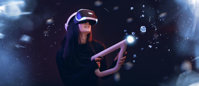 Beautiful woman in futuristic dress over dark blue magic light background. Gamer girl in glasses of virtual reality with controller in hands. Augmented reality, game, hobby concept. VR.