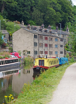 the rochdale canal running through hebden bridge with moored boats reflected in the water and stone buildings surrounded by trees