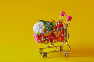 Grocery shopping cart with fresh vegetables and fruits. Isolated on yellow background. Free GMO....