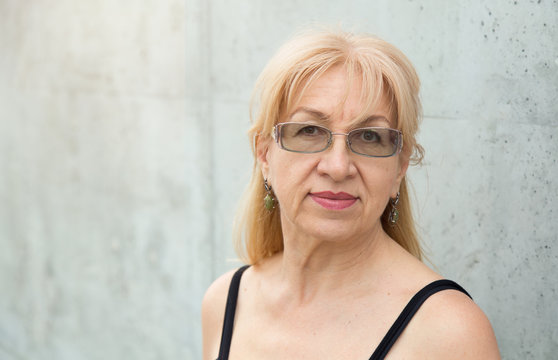 Close up portrait of happy mature woman in glasses, age 55 or 60 years against the gray wall.
