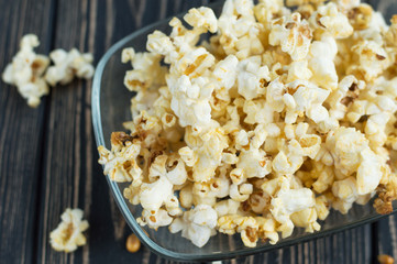Homemade vegan fresh crunchy salted popcorn in a glass plate on a dark wooden table. A useful snack for a movie, an idea for a meal. Dark mood close-up top view.