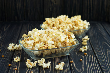 Homemade vegan fresh crunchy salted popcorn in a glass plate on a dark wooden table. A useful snack for a movie, an idea for a meal. Dark mood.