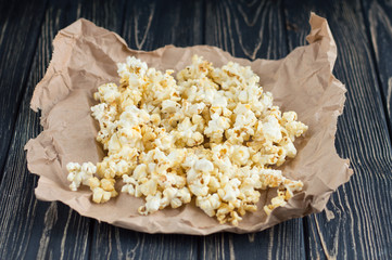 Homemade vegan fresh crunchy salted popcorn on brown craft paper lie on a dark wooden table. A useful snack for a movie, an idea for a meal. Dark mood top view.