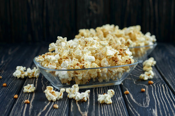 Homemade vegan fresh crunchy salted popcorn in a glass plate on a dark wooden table. A useful snack for a movie, an idea for a meal. Dark mood.