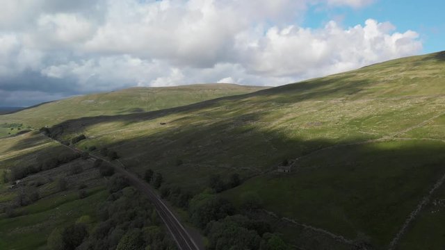 An aerial static shot looking at the Dent railway and the surrounding Cumbrian countryside. The scenery is green and sunny filmed in June. There are numerous clouds passing over the landscape.