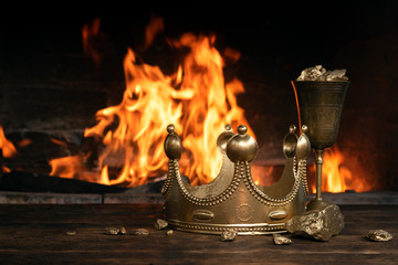 Golden crown and a goblet full of gold on the king table over burning fire background.